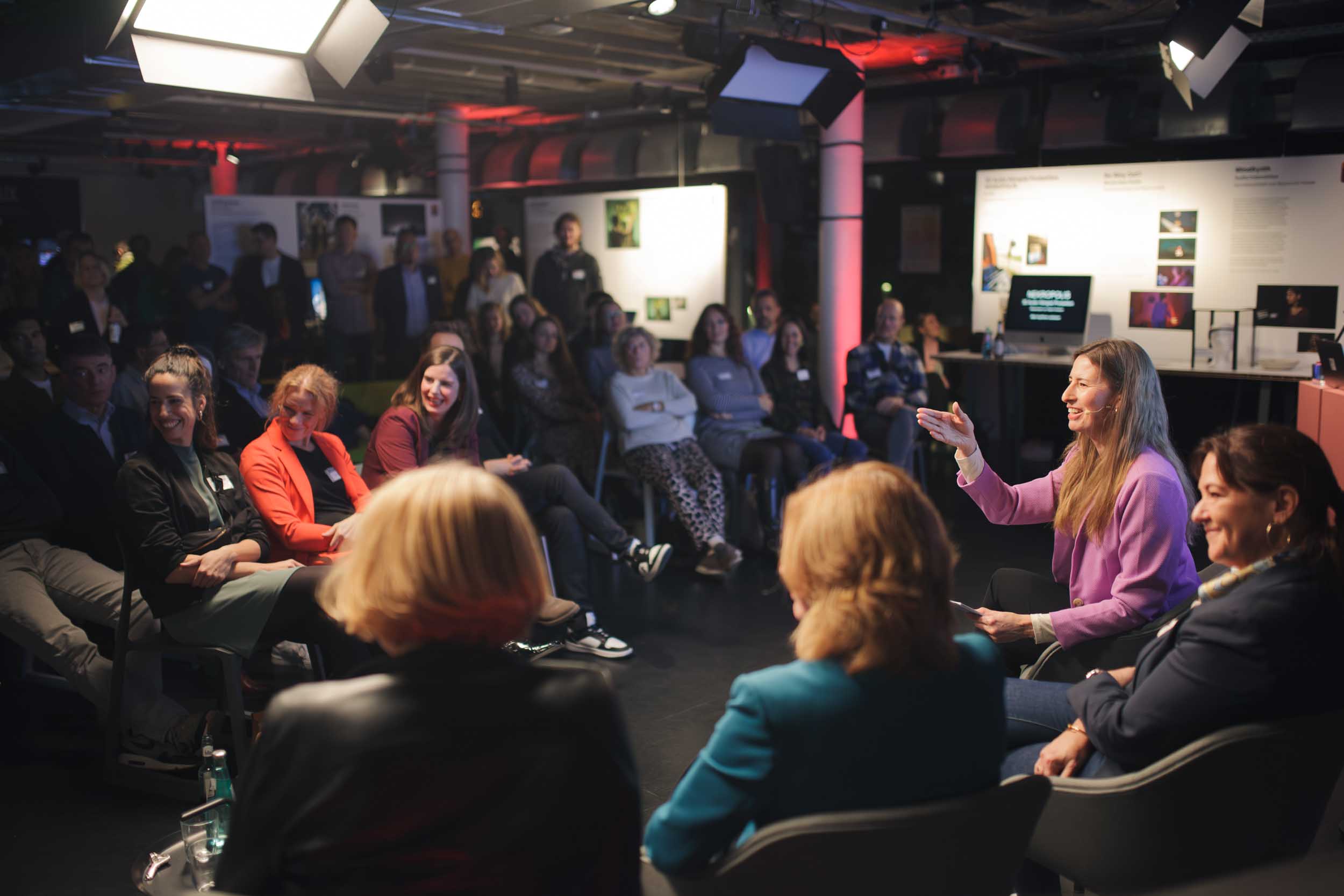 A woman with a pink jacket is sitting in front of a crowd. She is talking to someone of the crowd