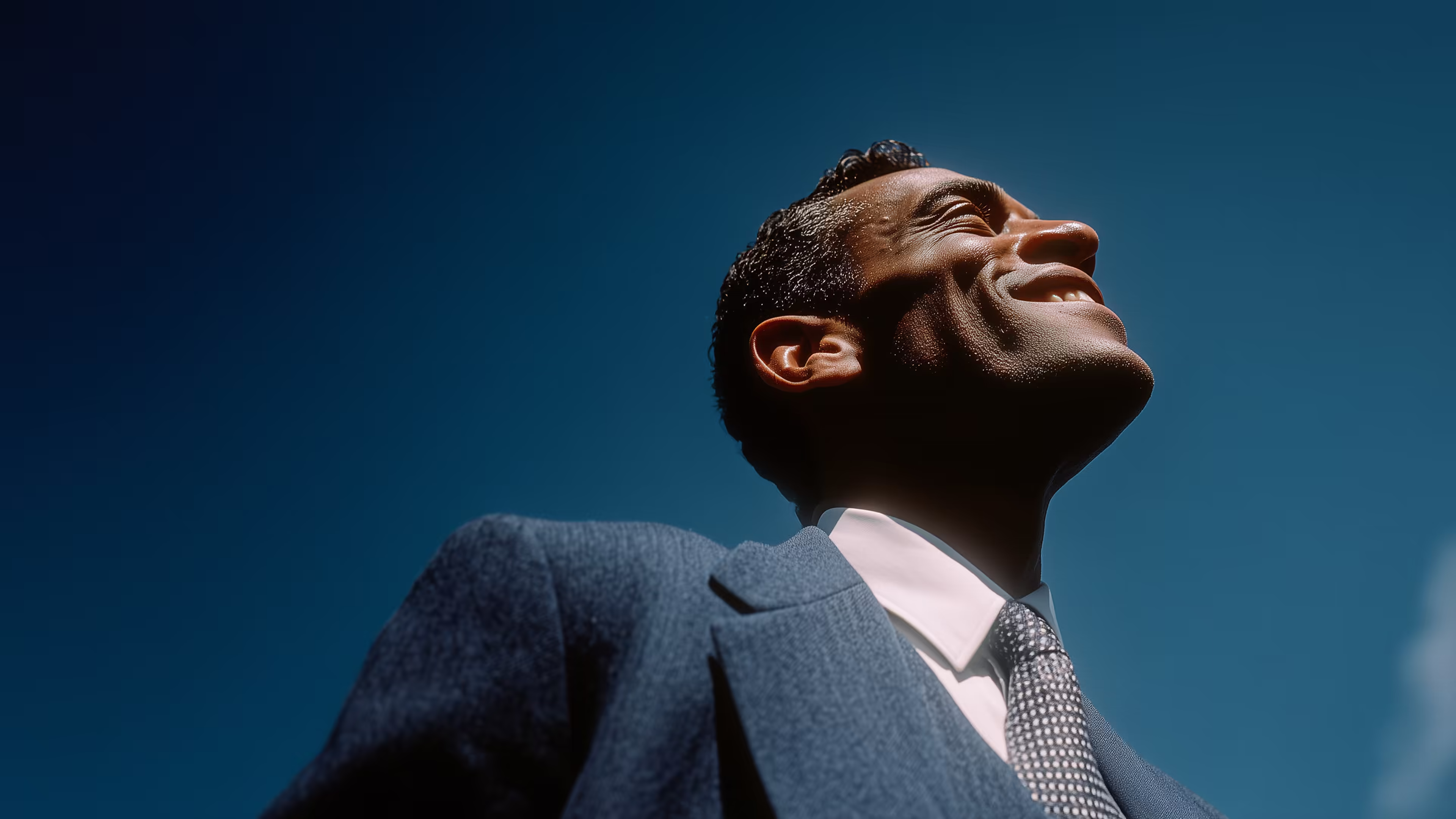Smiling man in a blue suit and patterned tie looking confidently upward against a clear blue sky.