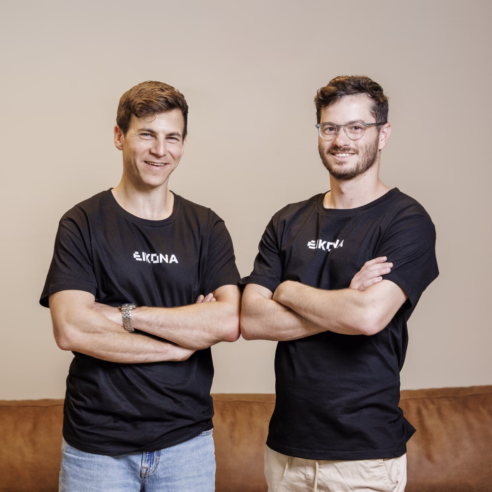 Two smiling men with crossed arms wearing black EIKONA t-shirts standing in front of a beige wall and brown couch.
