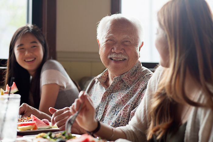 Smiling elderly man in patterned shirt dining with two young women. Bright, casual setting with food and drinks, mood is joyful and relaxed.