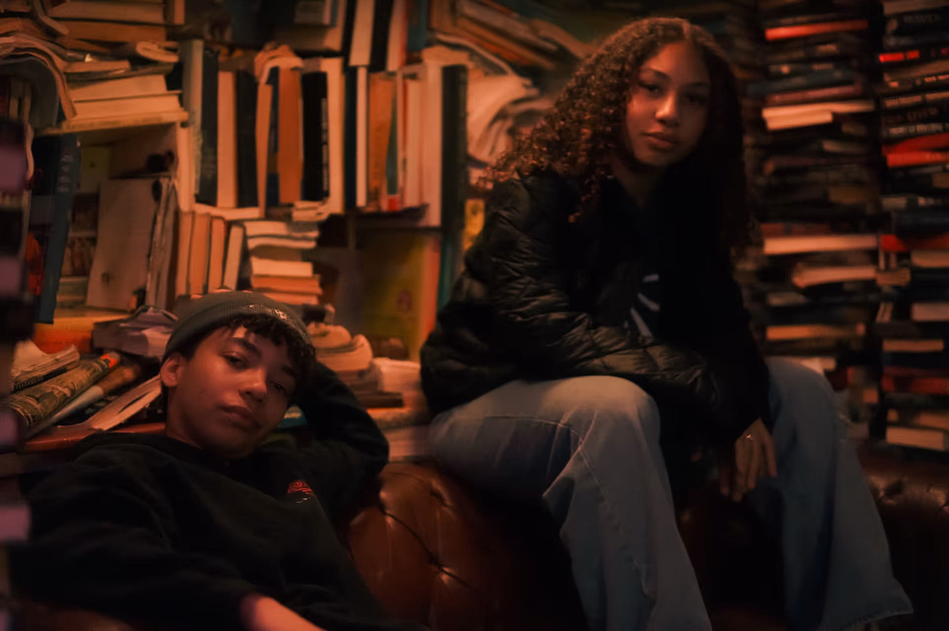 Two young people sitting on a leather couch surrounded by stacks of books in a cozy, dimly lit room.