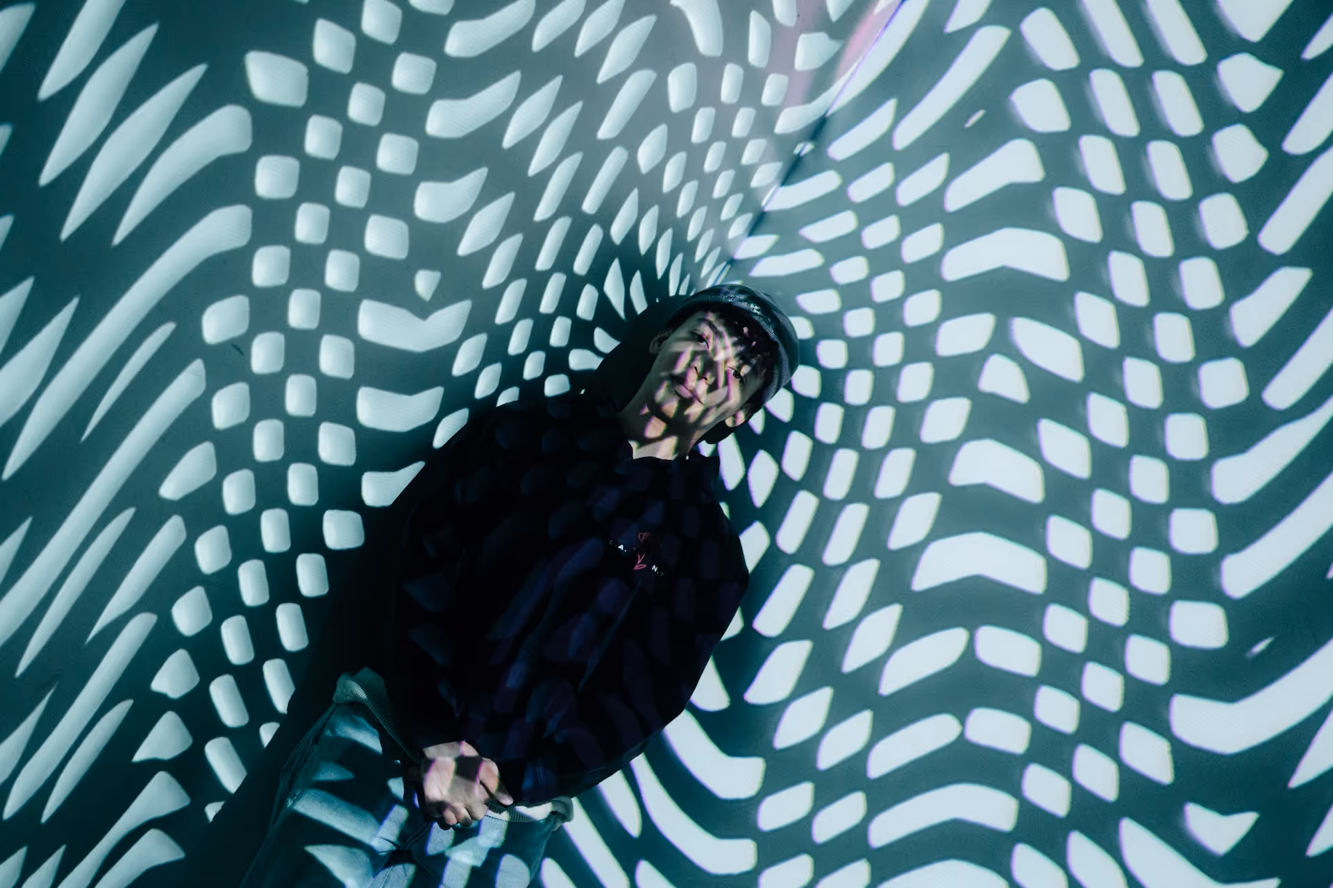 Person wearing a cap and dark clothing standing against a wall with abstract black and white geometric light patterns projected onto them.