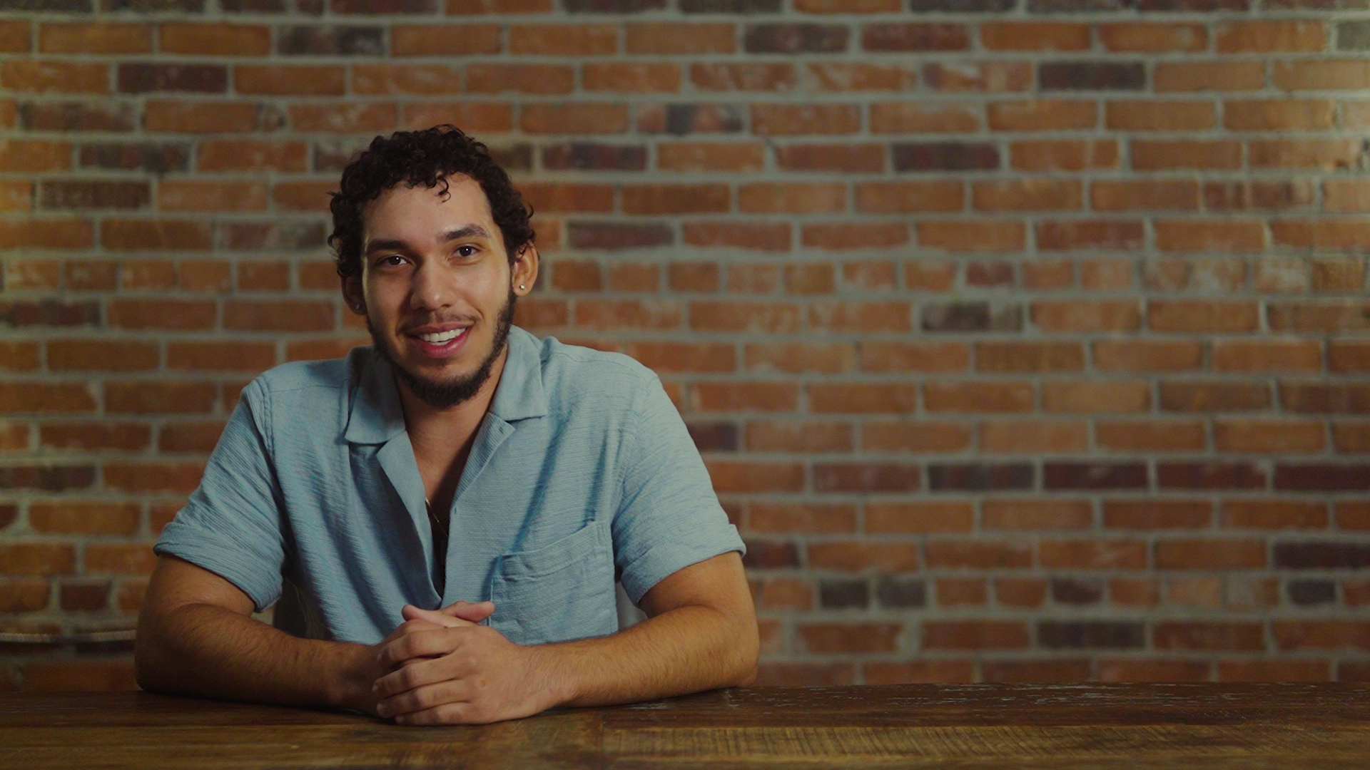 Young man with curly hair and beard sitting at a wooden table in front of a brick wall, smiling.