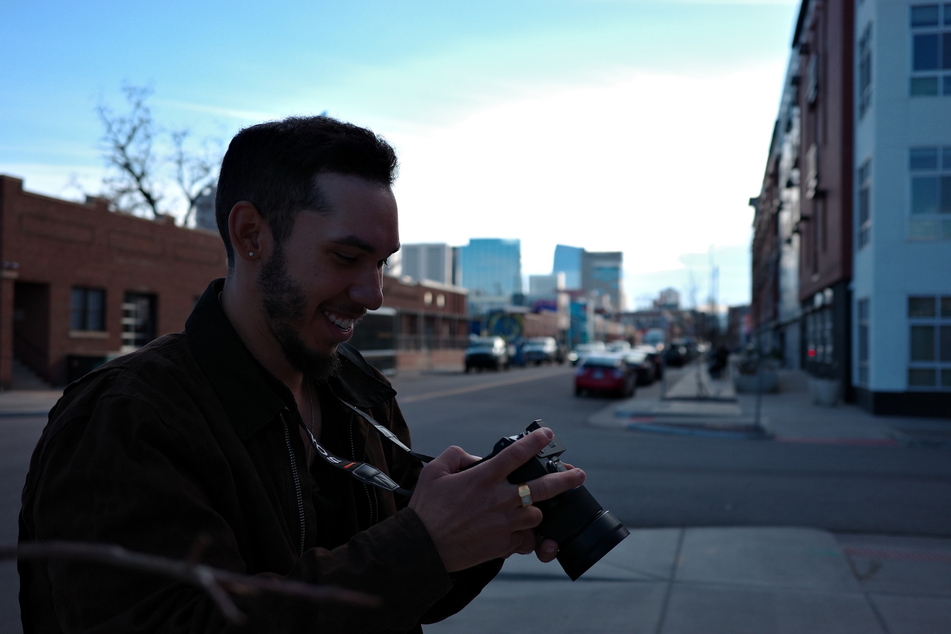 Smiling man holding and looking at a camera on a city street with buildings and cars in the background.