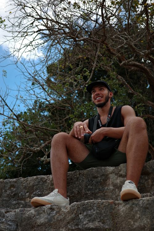 Smiling young man in a black tank top and cap sitting on stone steps holding a camera, with trees and blue sky behind him.