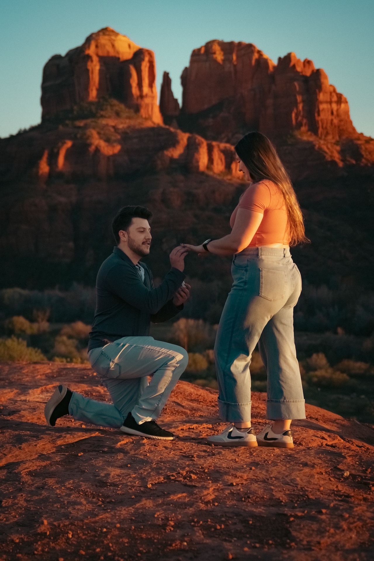 Man kneeling and proposing to woman on rocky terrain with large red rock formations in the background at sunset.