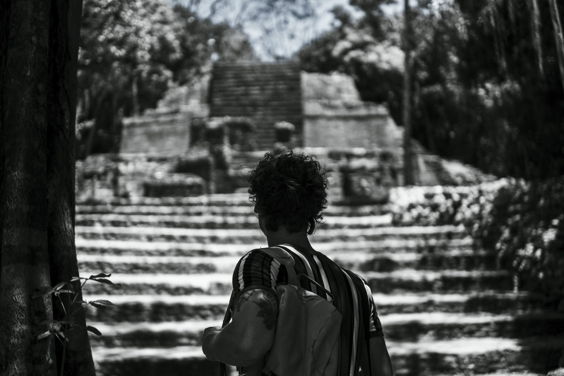 Man with backpack facing and looking up at an ancient stone pyramid or temple with steps surrounded by trees.