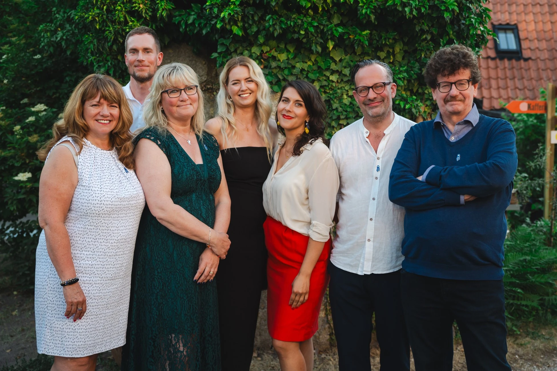 Seven adults standing outdoors in front of green foliage, smiling at the camera; five women and two men dressed in semi-formal attire.