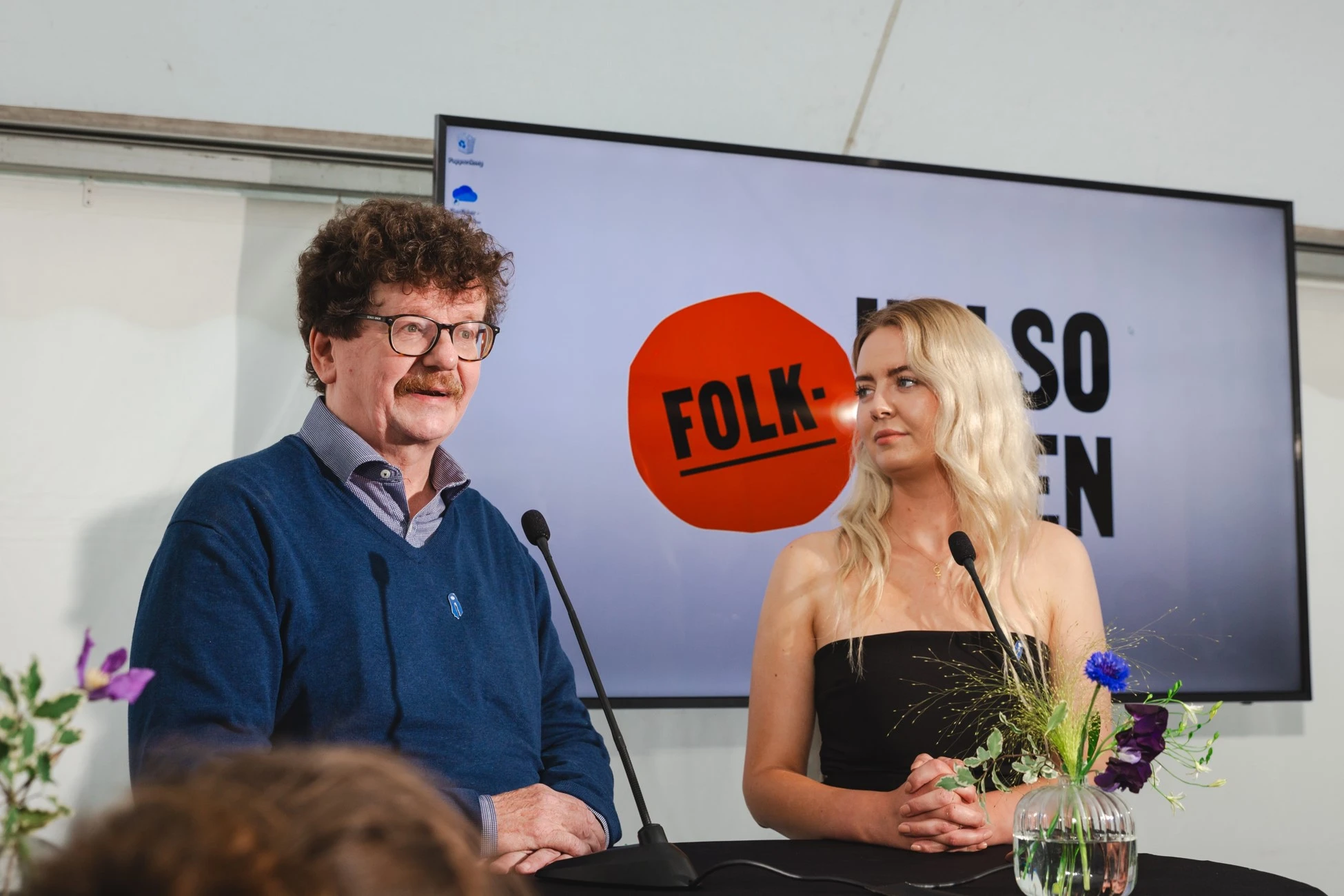 Man with curly hair and glasses speaking at a microphone beside a woman