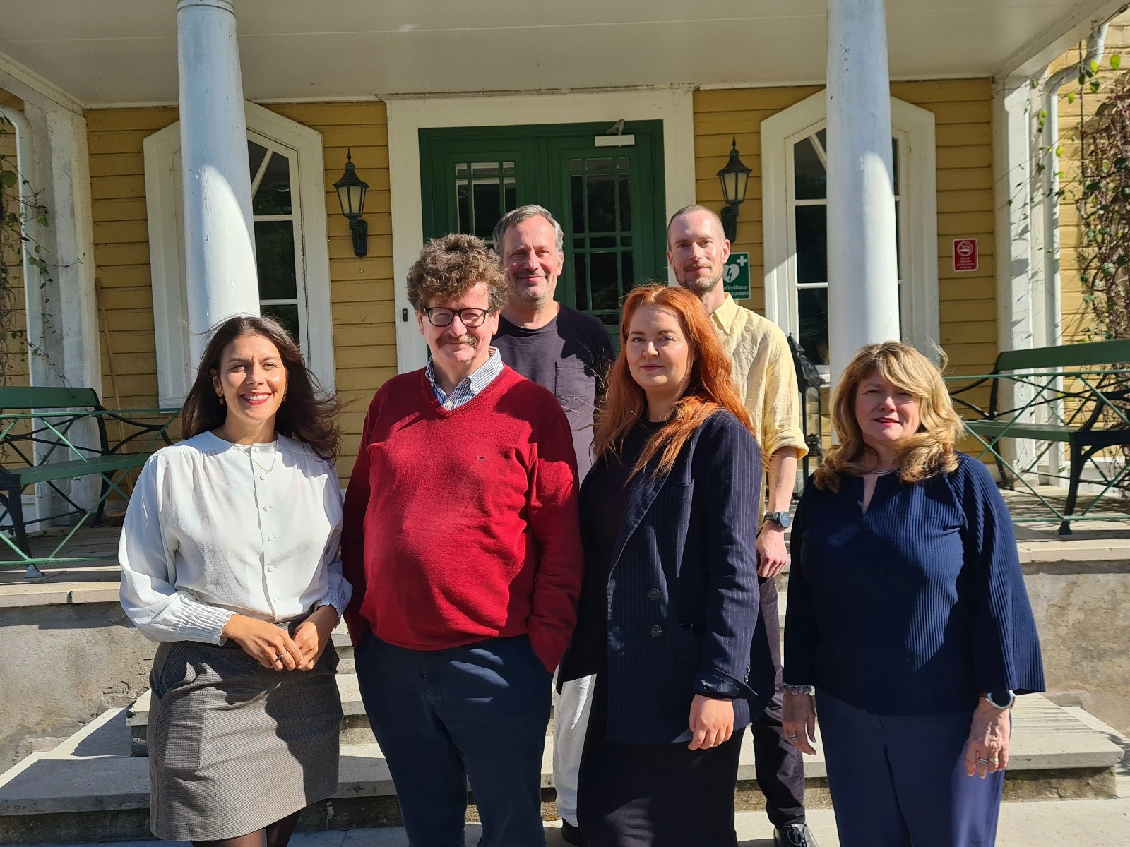Group of six adults standing outside a yellow building with green double doors, smiling at the camera in sunlight.