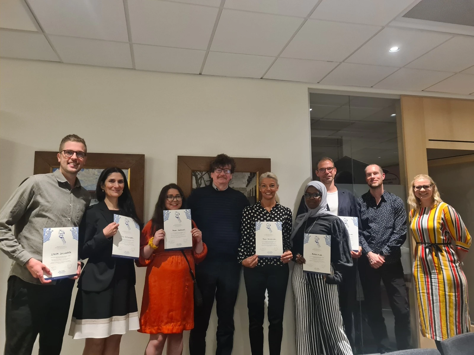Group of nine people standing indoors, six of them holding certificates and smiling at the camera.