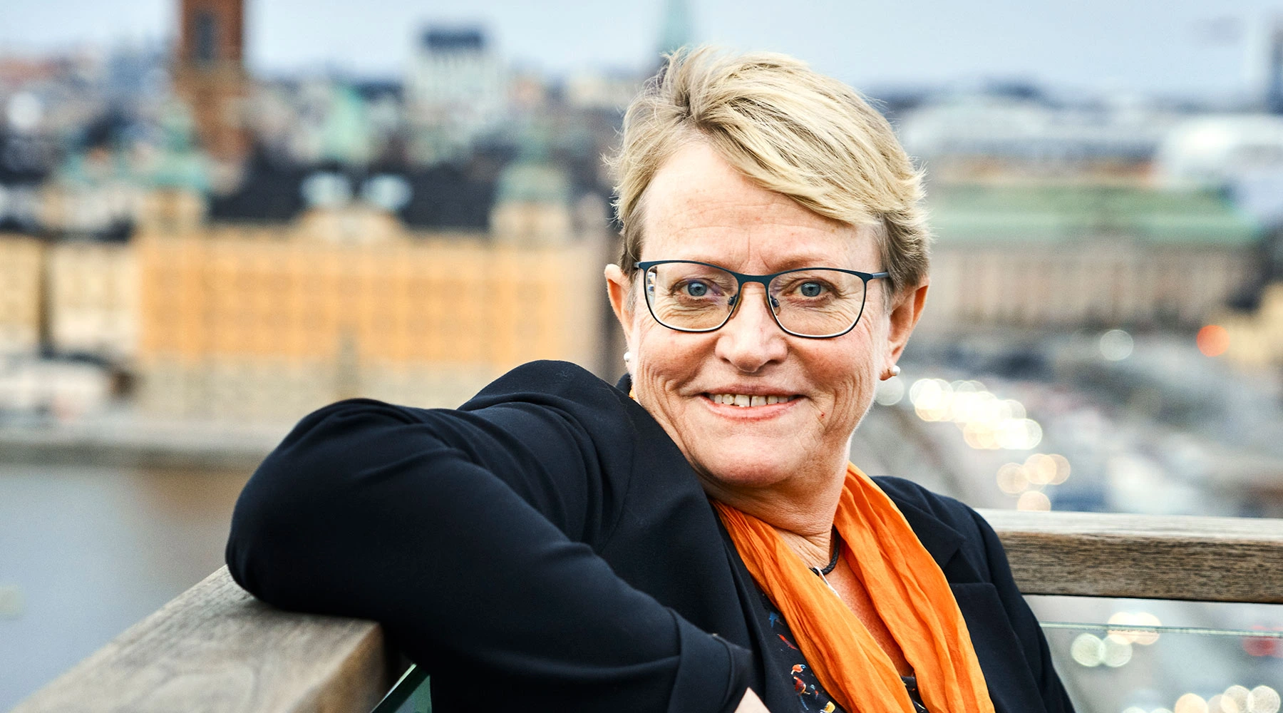 Smiling woman with short blonde hair, glasses, and an orange scarf leaning on a wooden railing with a blurred cityscape background.