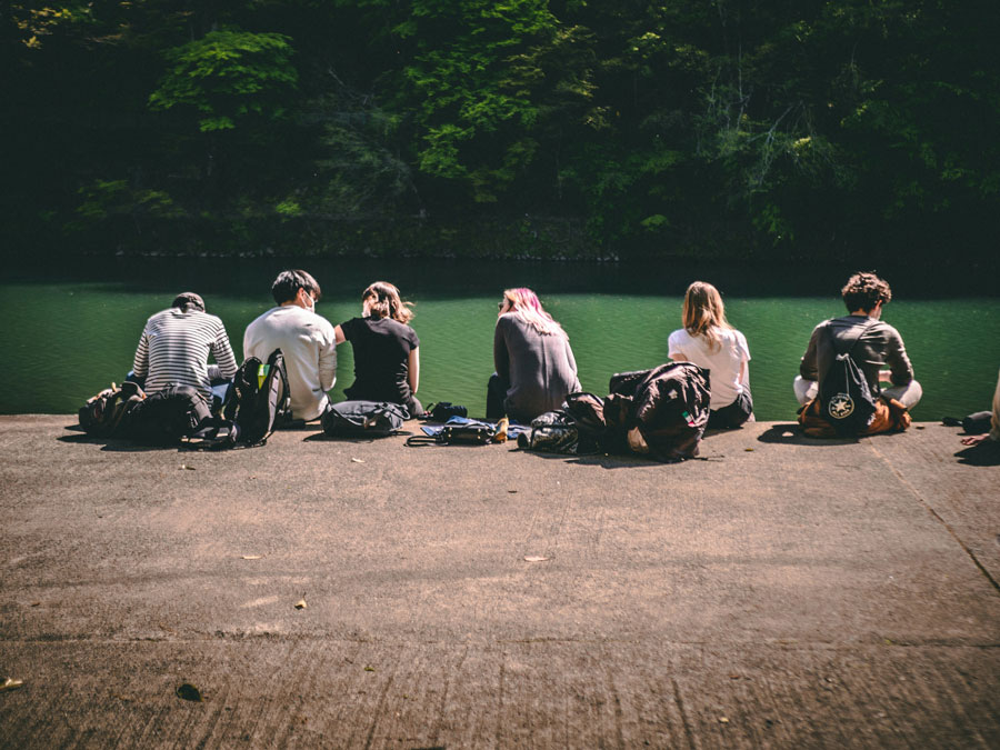 Six people sit on a concrete ledge by a calm green lake, surrounded by trees and backpacks, conveying a peaceful, relaxed setting.
