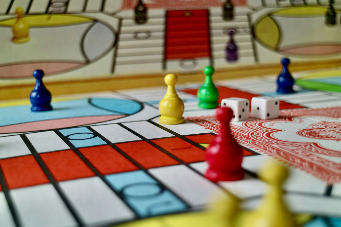 Colorful board game with yellow, red, blue, and green player pieces on a patterned surface. Two white dice are rolled mid-game, creating a lively atmosphere.