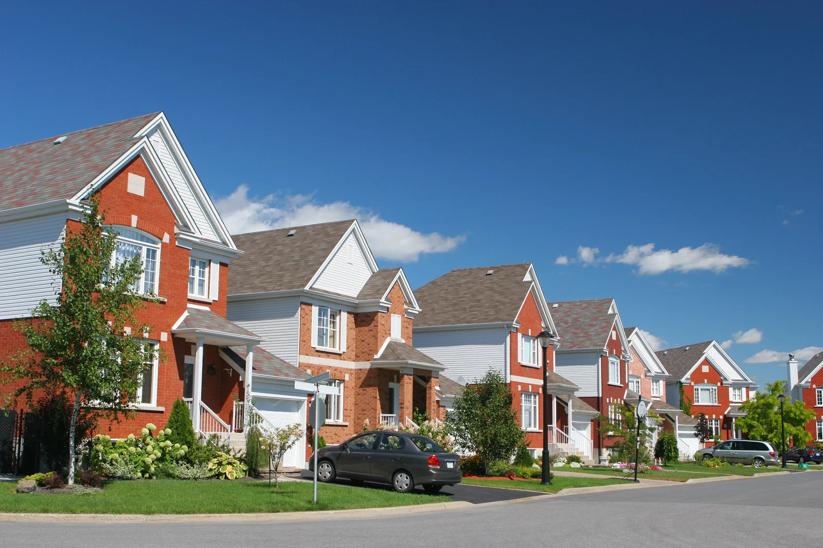 Row of suburban two-story houses with brick and white siding under a blue sky, with cars parked in driveways.