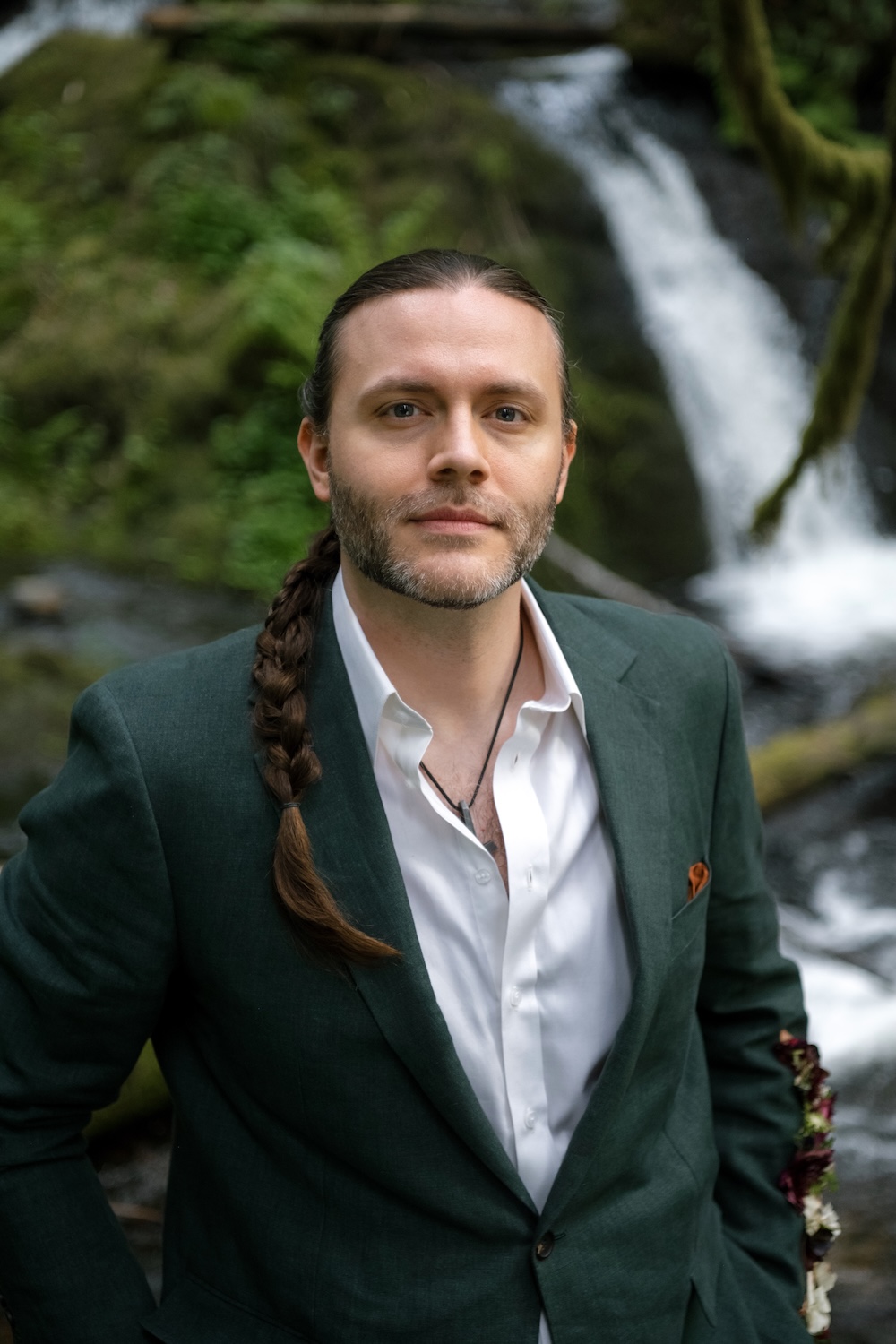 A man with long braided hair and a green jacket stands in front of a waterfall and mossy rocks, looking calm and confident.