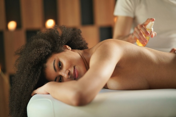 A woman smiles gently while lying on a massage table as a therapist applies massage oil in a warmly lit space.