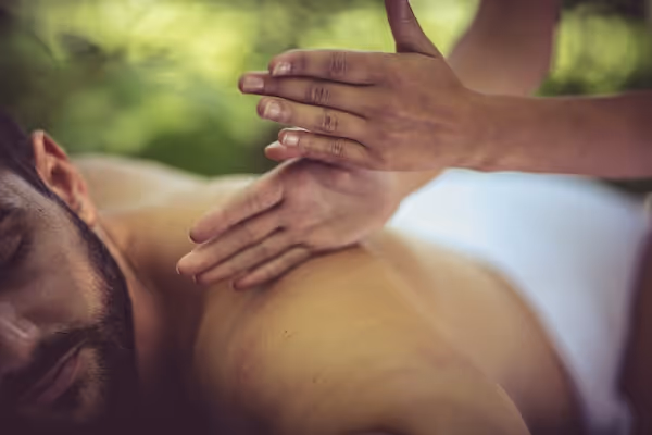 A man relaxes during a massage, receiving rhythmic hand techniques on his back in a natural outdoor setting.