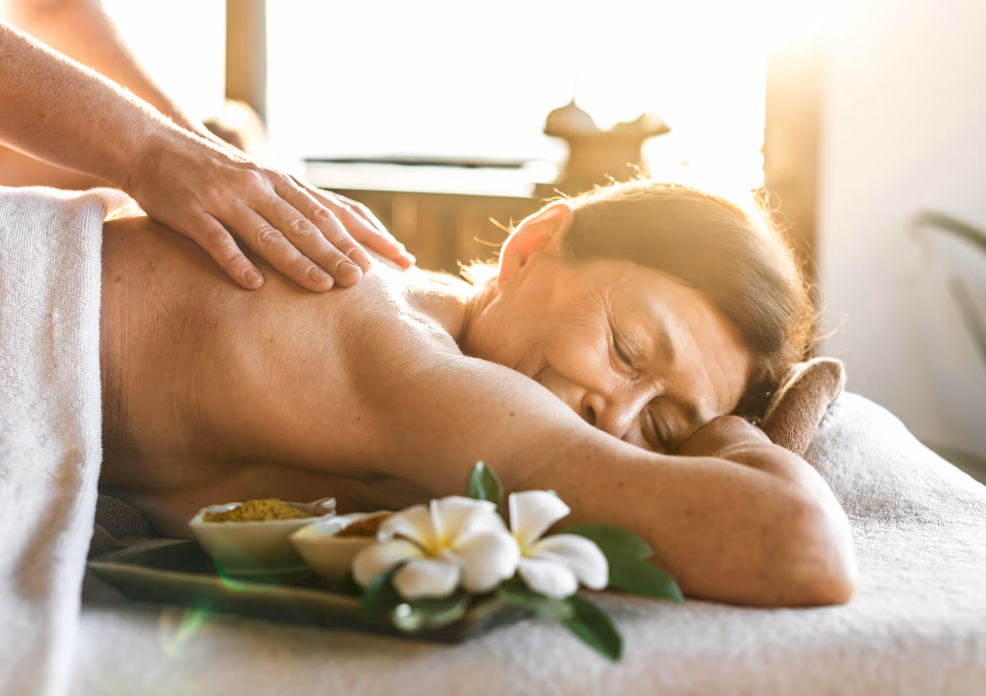 An older woman lies peacefully on a massage table, smiling with her eyes closed as a massage therapist gently presses on her shoulder. A soft towel, flowers, and natural bowls of ingredients rest nearby, with warm sunlight streaming into the room.