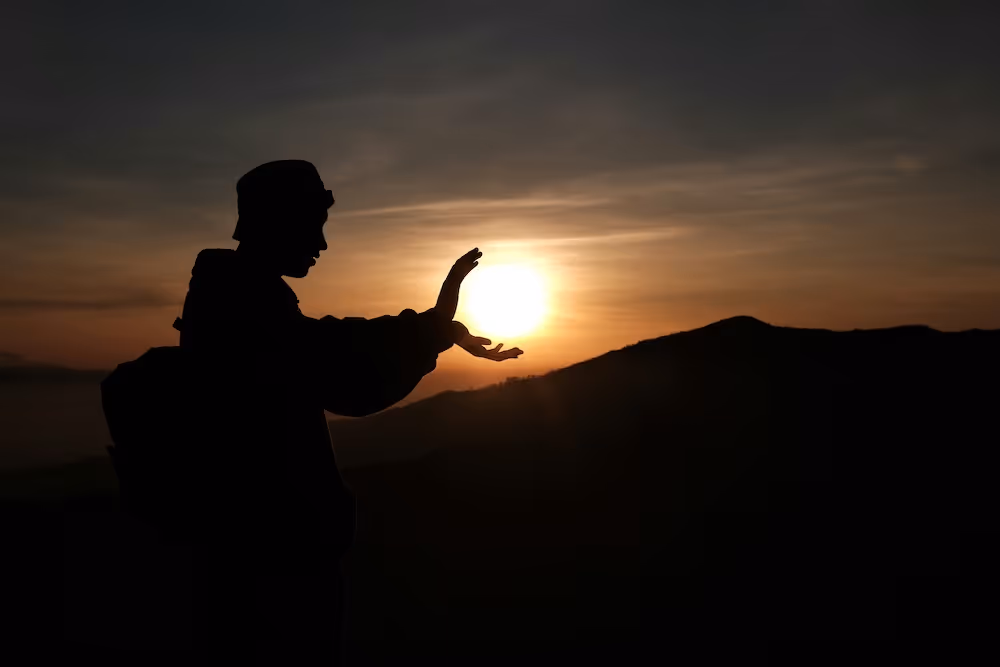 A person’s silhouette on a mountain ridge holds their hands around the setting sun, creating the impression of cradling light.