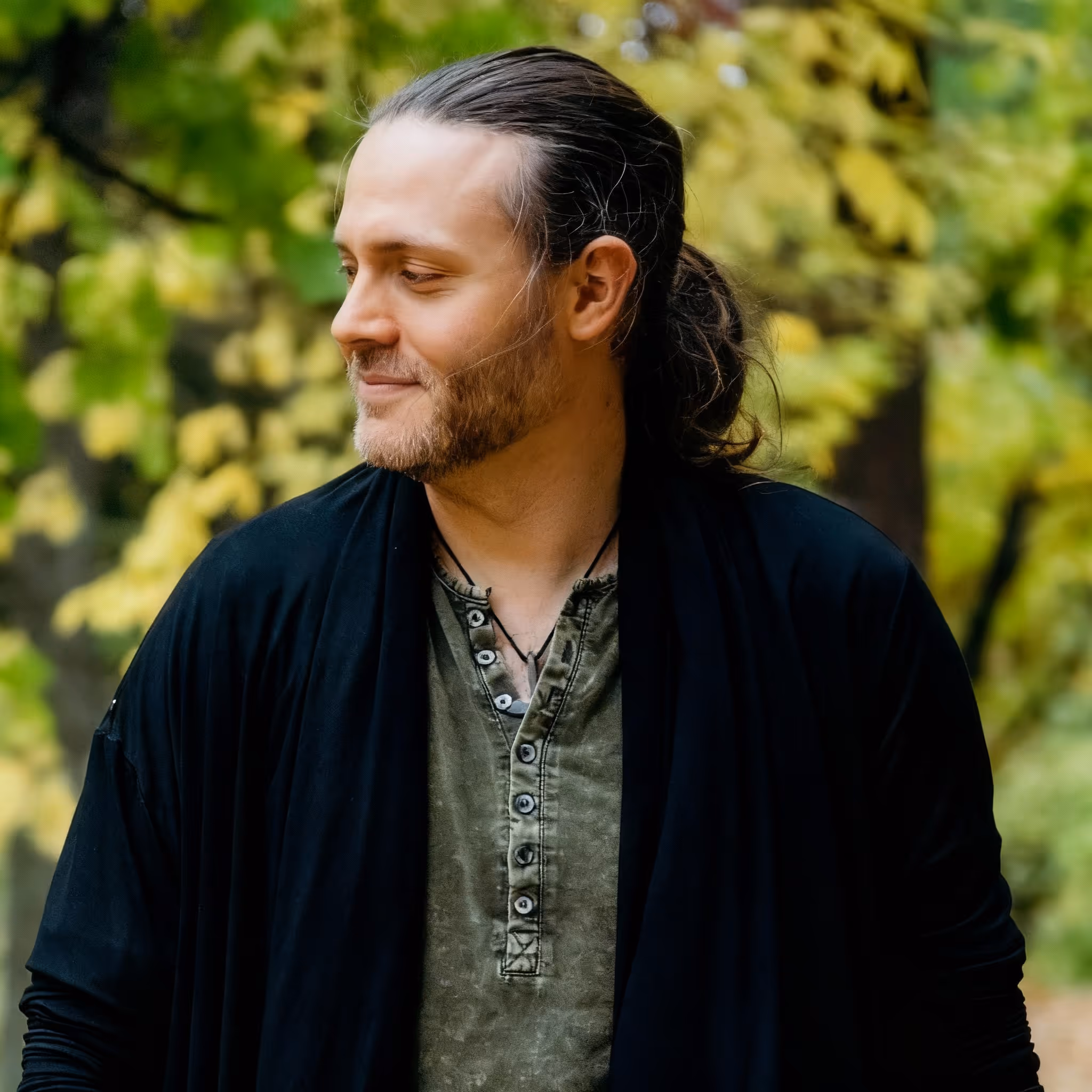 Forest stands outdoors in a forest setting, wearing a dark green shirt and black cardigan, looking slightly to the side with a soft smile. The background is filled with warm green and golden leaves, softly blurred for a natural, serene effect.