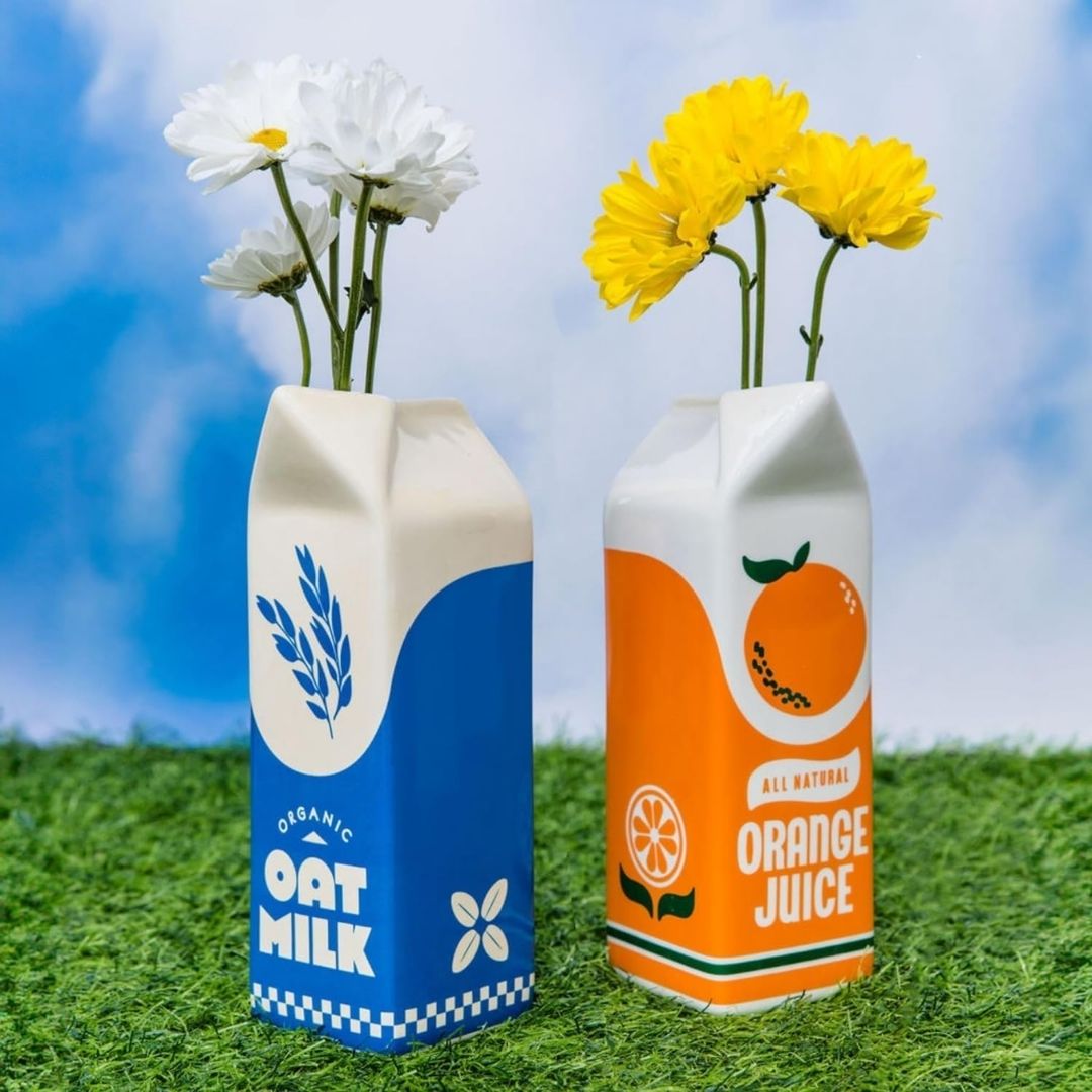A ceramic vase shaped like a retro orange juice carton with a vintage design and bright, colorful details, displayed on a counter.