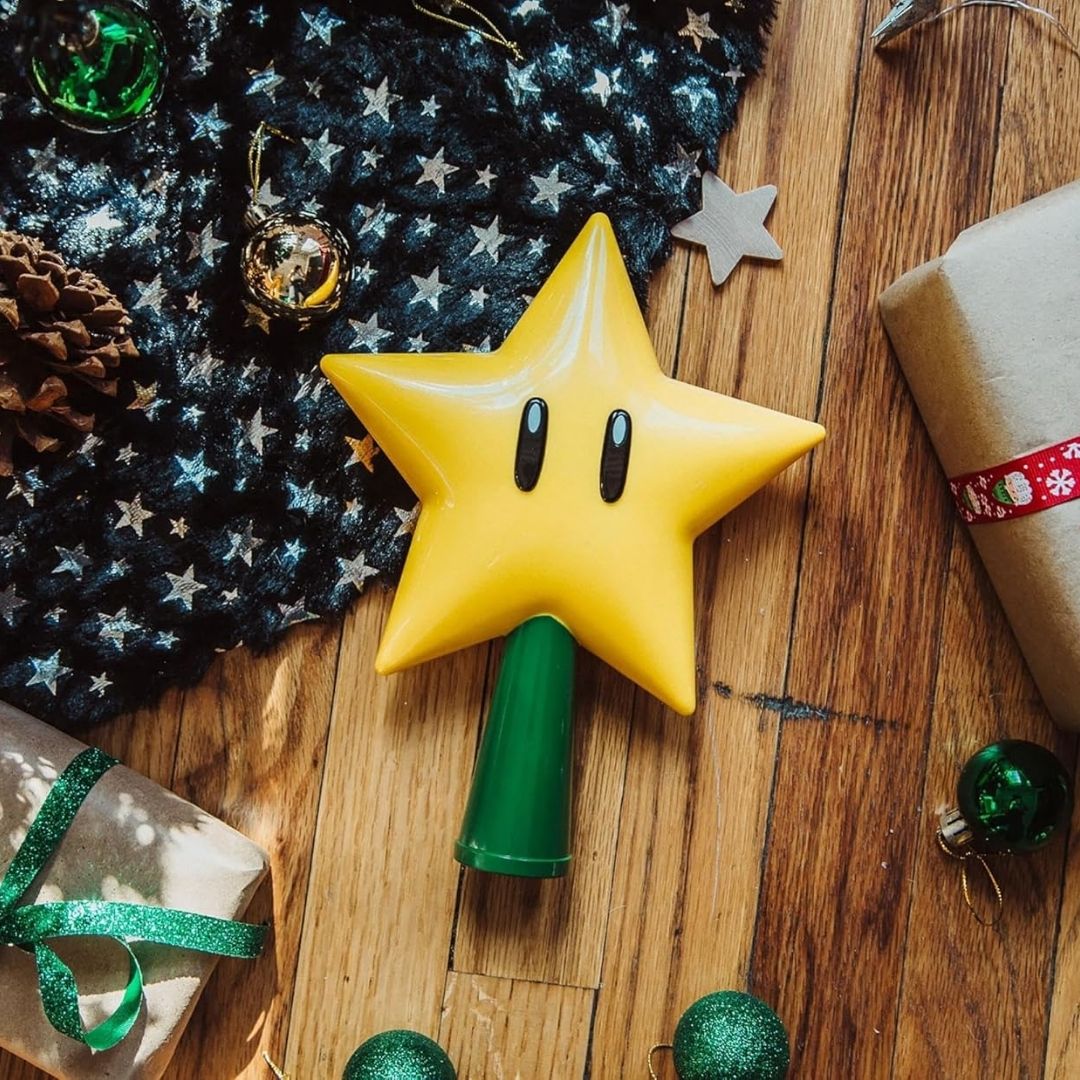 A Super Mario Light-Up Star Tree Topper, shaped like a bright yellow star with black eyes, glows atop a holiday tree.