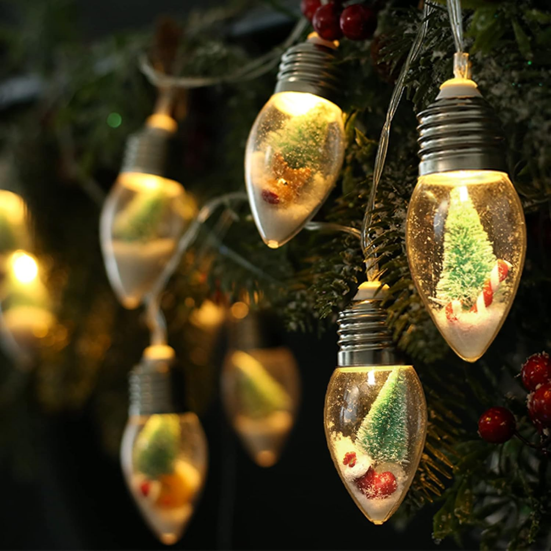 A string of globe-shaped Christmas lights with clear bulbs, each filled with faux pine, snow, and festive ornaments, displayed as cozy indoor holiday decor.