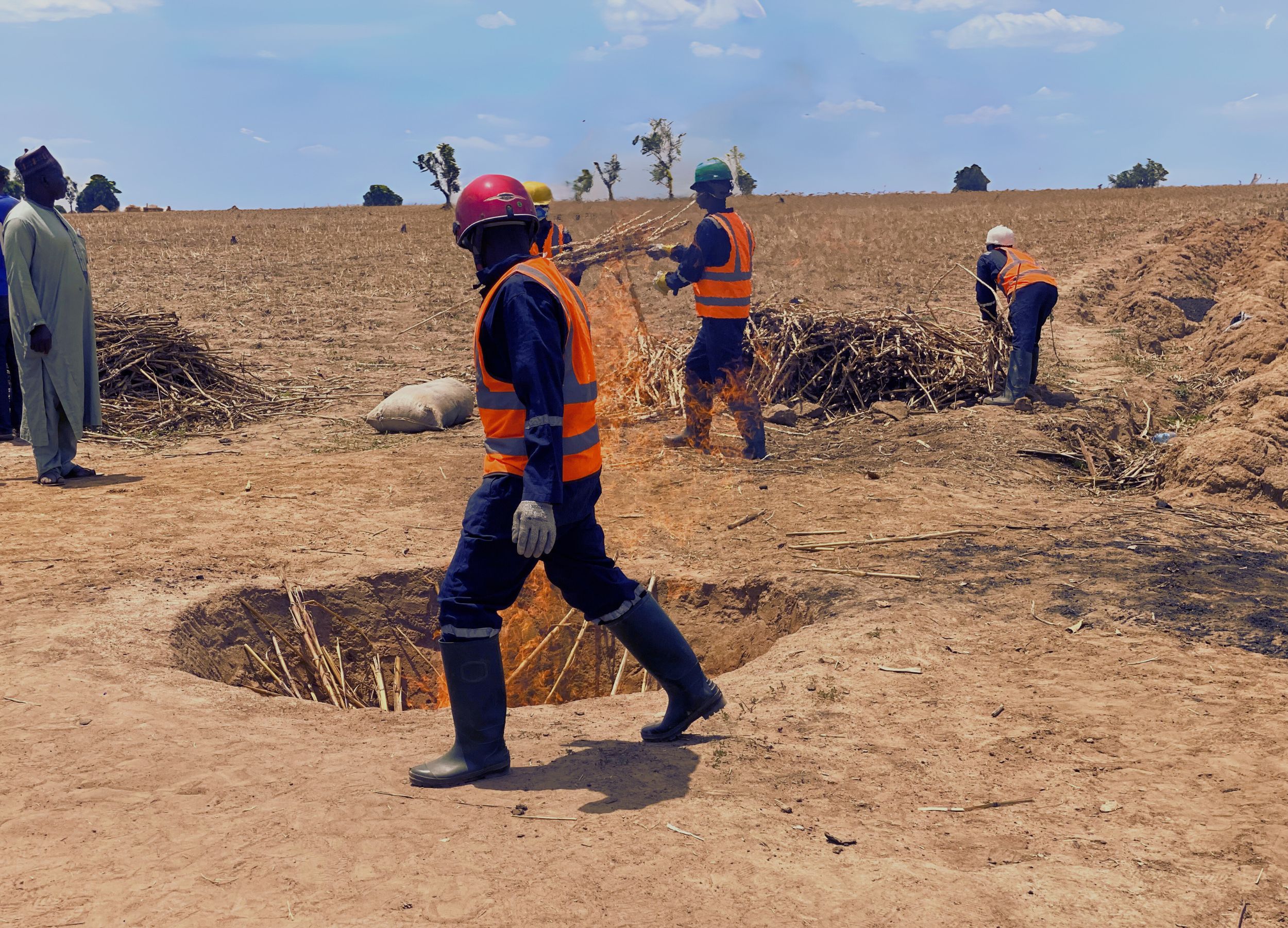Workers in safety helmets and orange vests burning dry branches in a large hole in a dry, open field under a blue sky.