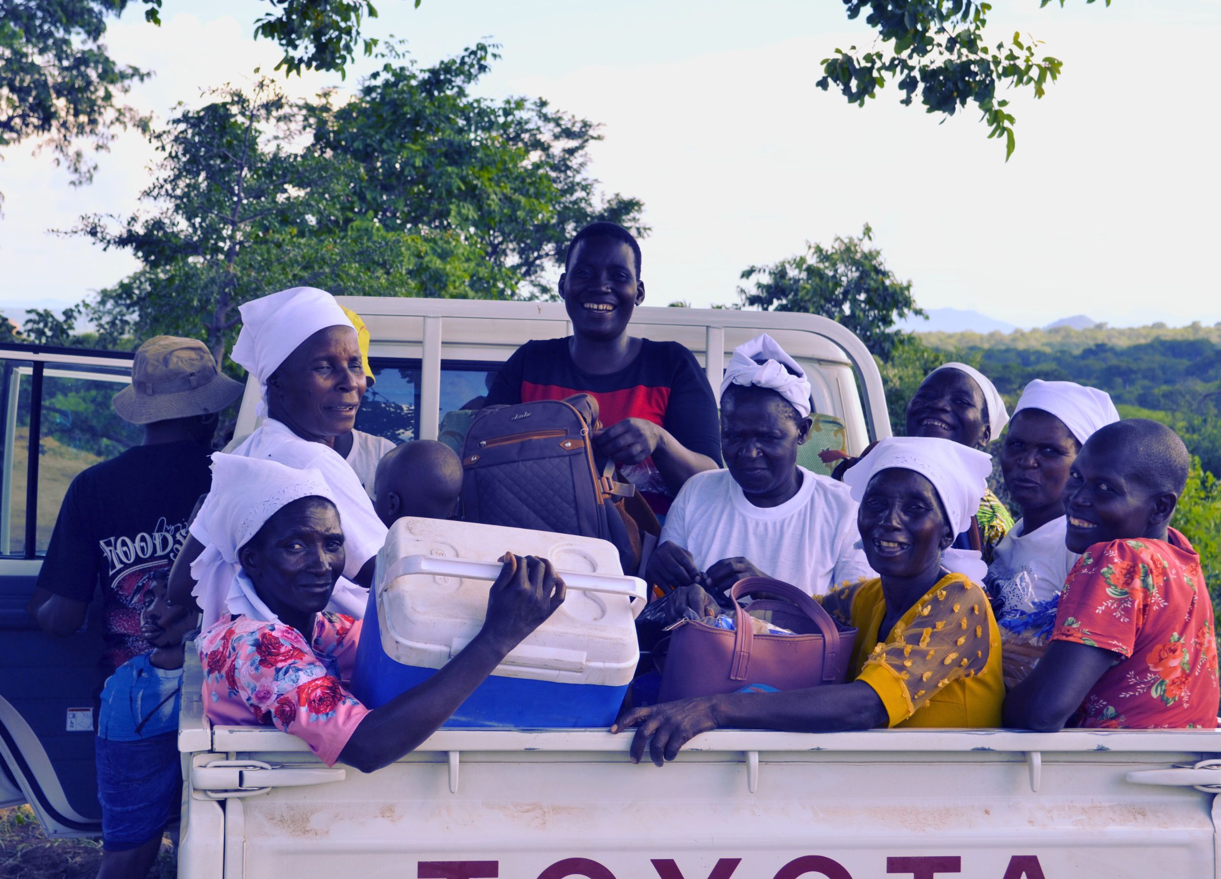 A group of smiling people, mostly women wearing headscarves, sitting in the back of a white Toyota pickup truck in a rural outdoor setting.