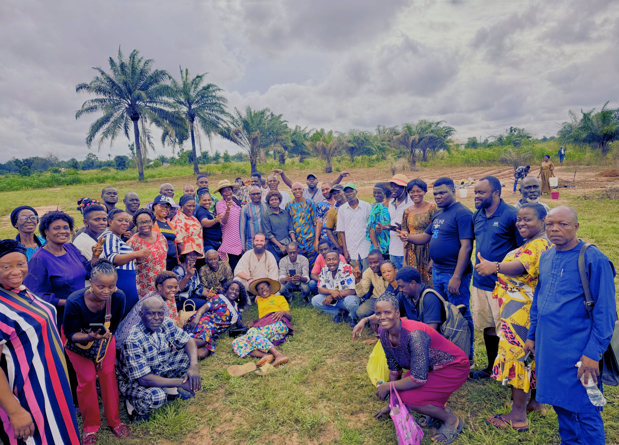 Large group of diverse adults posing together outdoors on grass with palm trees and cloudy sky in the background.