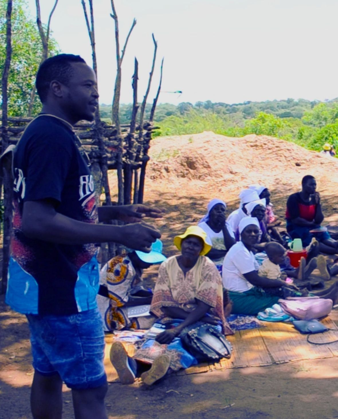 Man standing and speaking to a group of seated women and a child outdoors on a mat in a rural setting with trees and a dirt mound in the background.