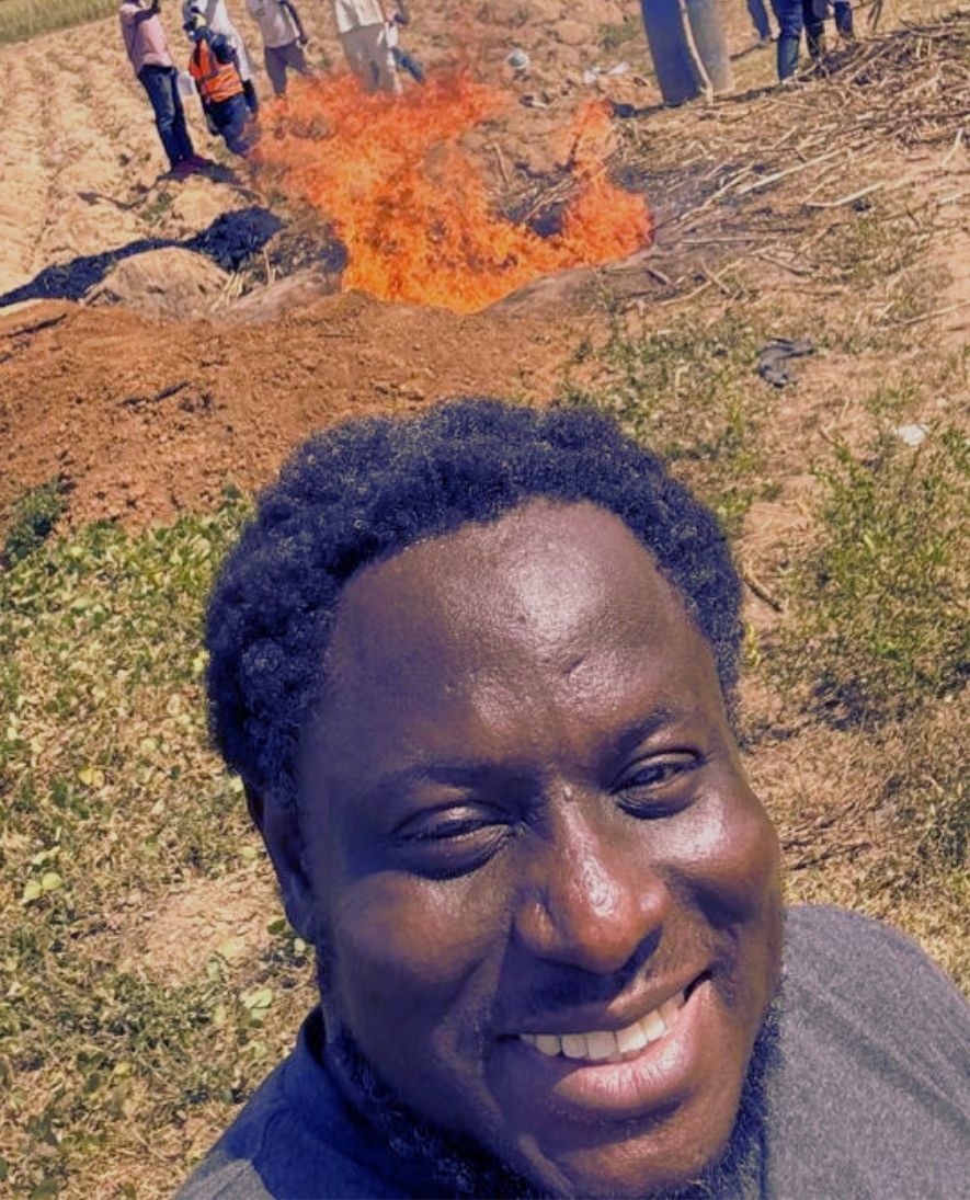 Smiling man taking a selfie outdoors with a fire burning in the background and a group of people standing behind it.