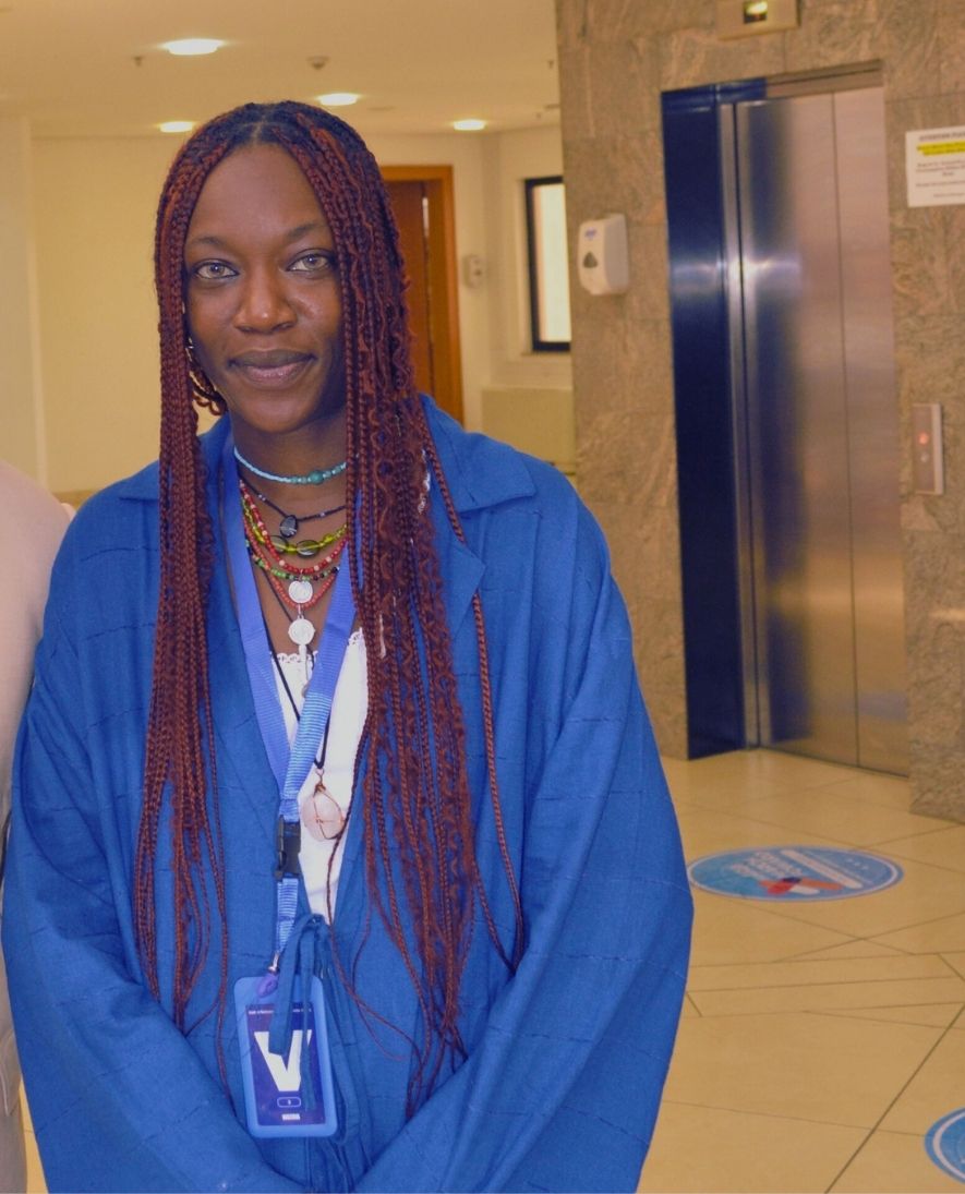 Person with long braided hair wearing a blue jacket and colorful necklaces standing indoors near an elevator.
