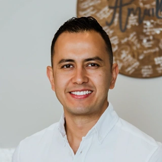Portrait of a smiling man with short dark hair wearing a white button-up shirt in front of a blurred background.