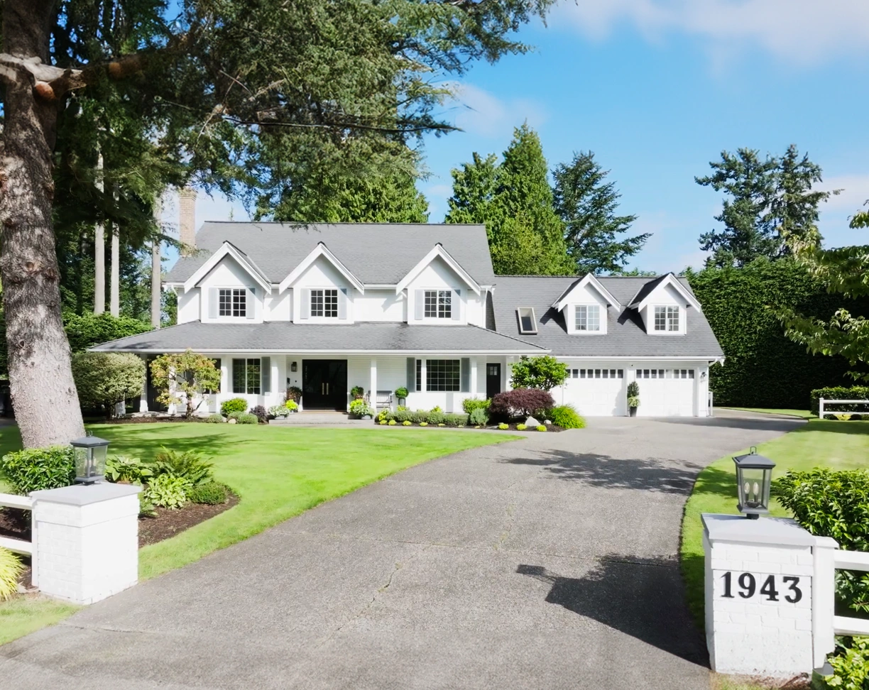 Large white two-story house with gray roof, a three-car garage, neatly trimmed green lawn, and a driveway leading to the house with the number 1943 on a white pillar.