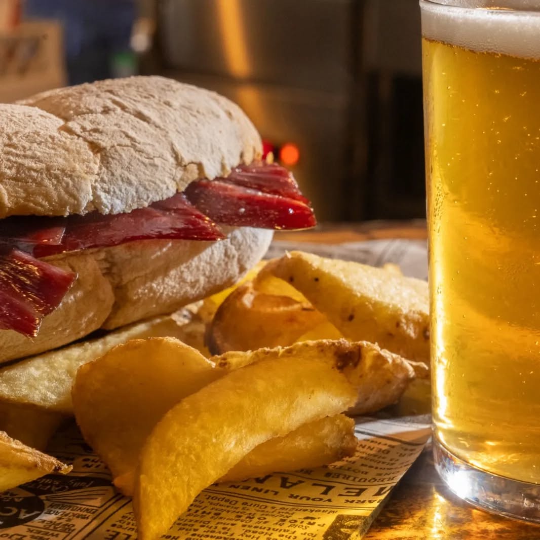 Bocadillo de jamón con patatas fritas y vaso de cerveza dorada en mesa de madera.