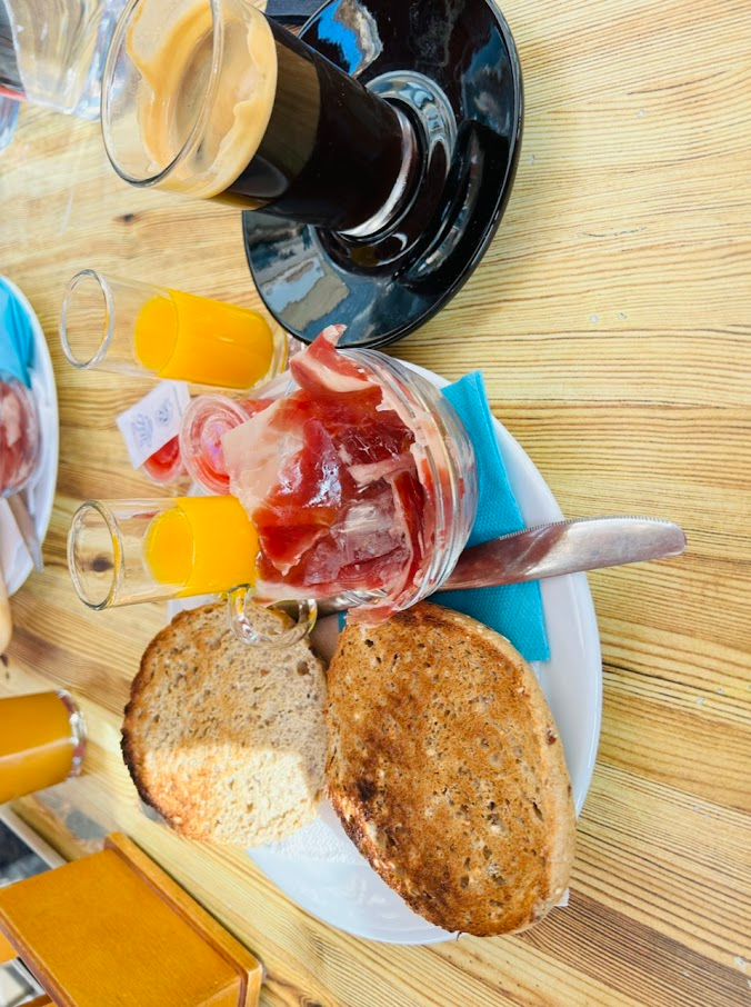 Desayuno con café en vaso, dos vasos de jugo de naranja, pan tostado y jamón serrano en un frasco