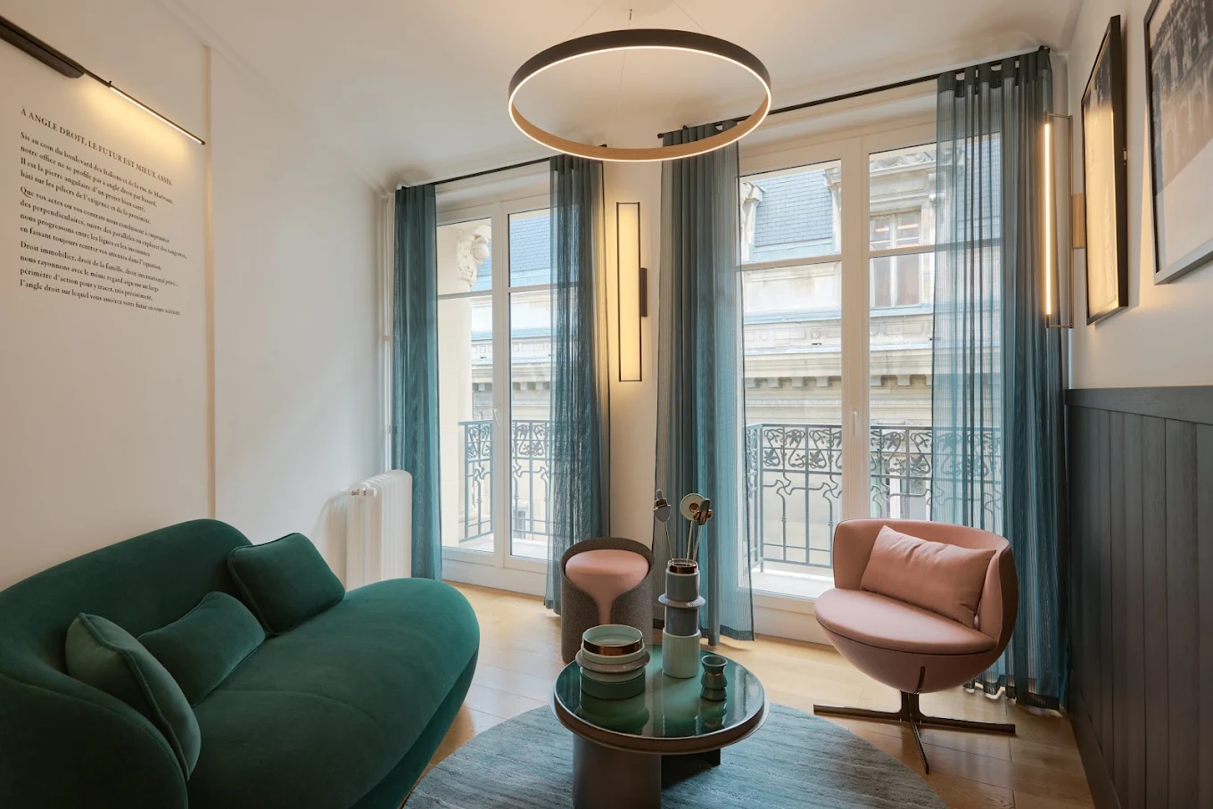 Modern living room with a green sofa, pink armchair, round coffee table, blue sheer curtains, and large windows overlooking a classic building facade.