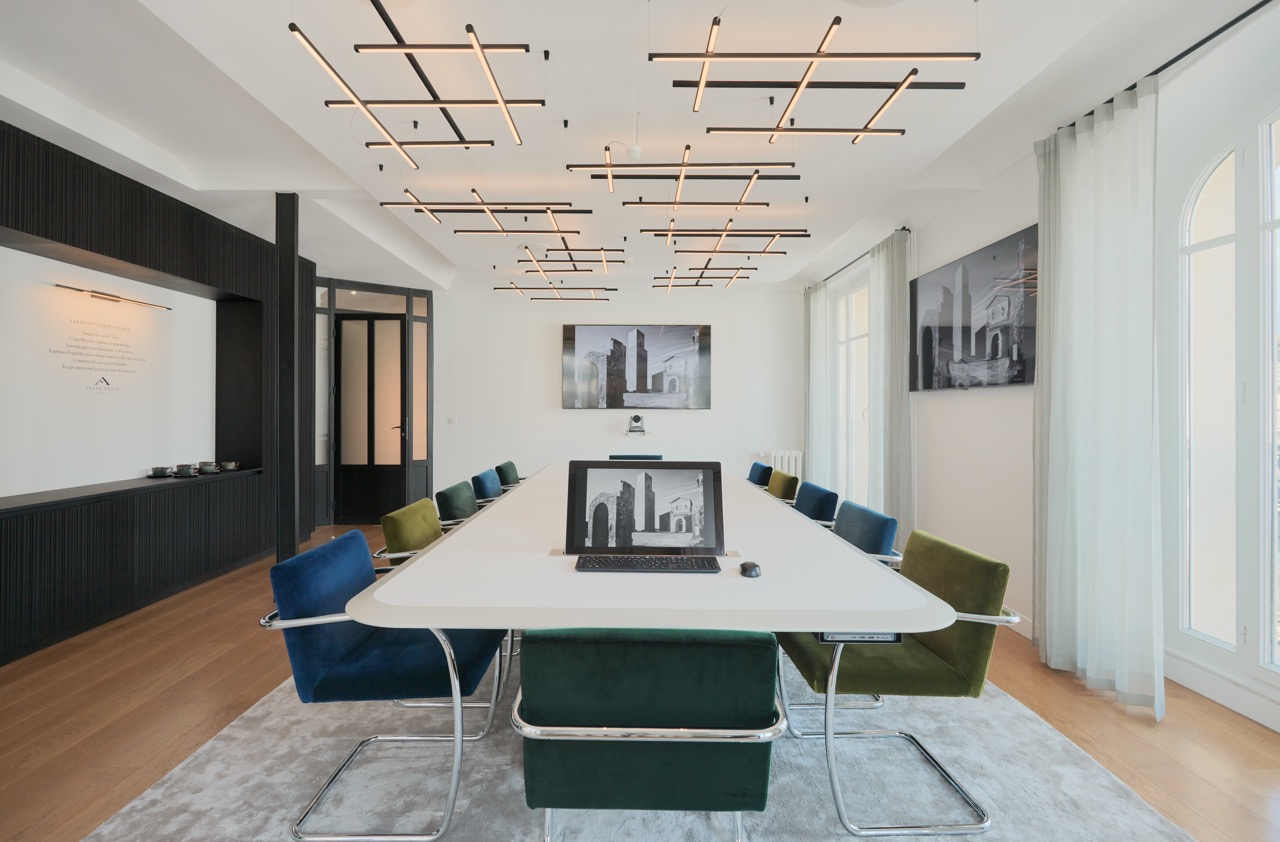 Modern conference room with a long white table, colorful velvet chairs, abstract ceiling lights, and black and white architectural photos on walls.