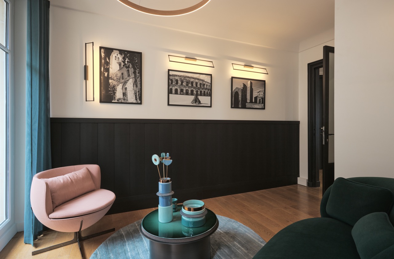 Modern living room with a pink armchair, dark green sofa, round table with decorative vases, blue curtain, wooden floor, and black-and-white framed photos on a white wall.