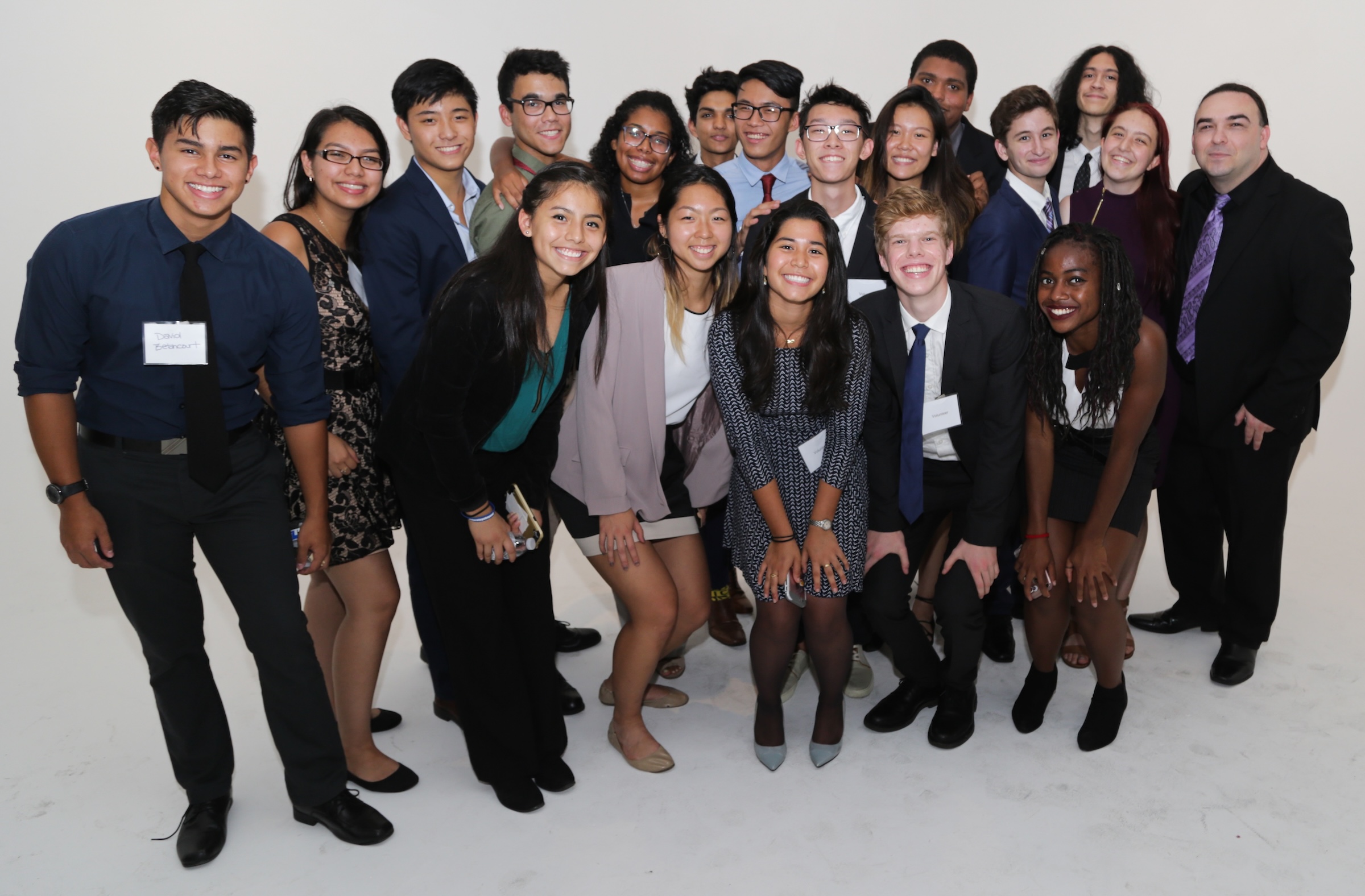 Group portrait of smiling young adults dressed in formal and semi-formal attire against a plain white background.