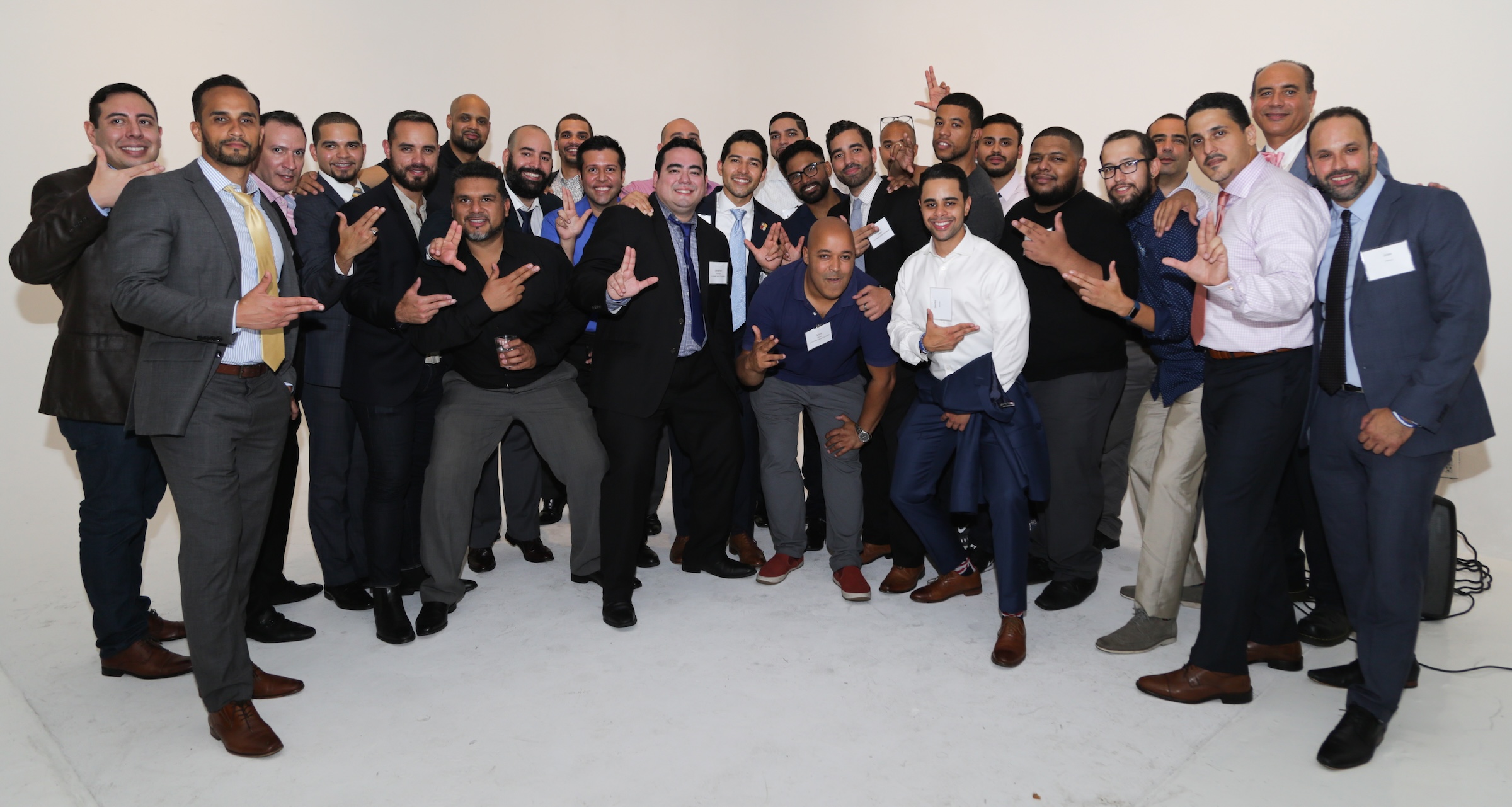 Group of men dressed in business casual and formal wear posing indoors, many making hand gestures resembling the letter L.