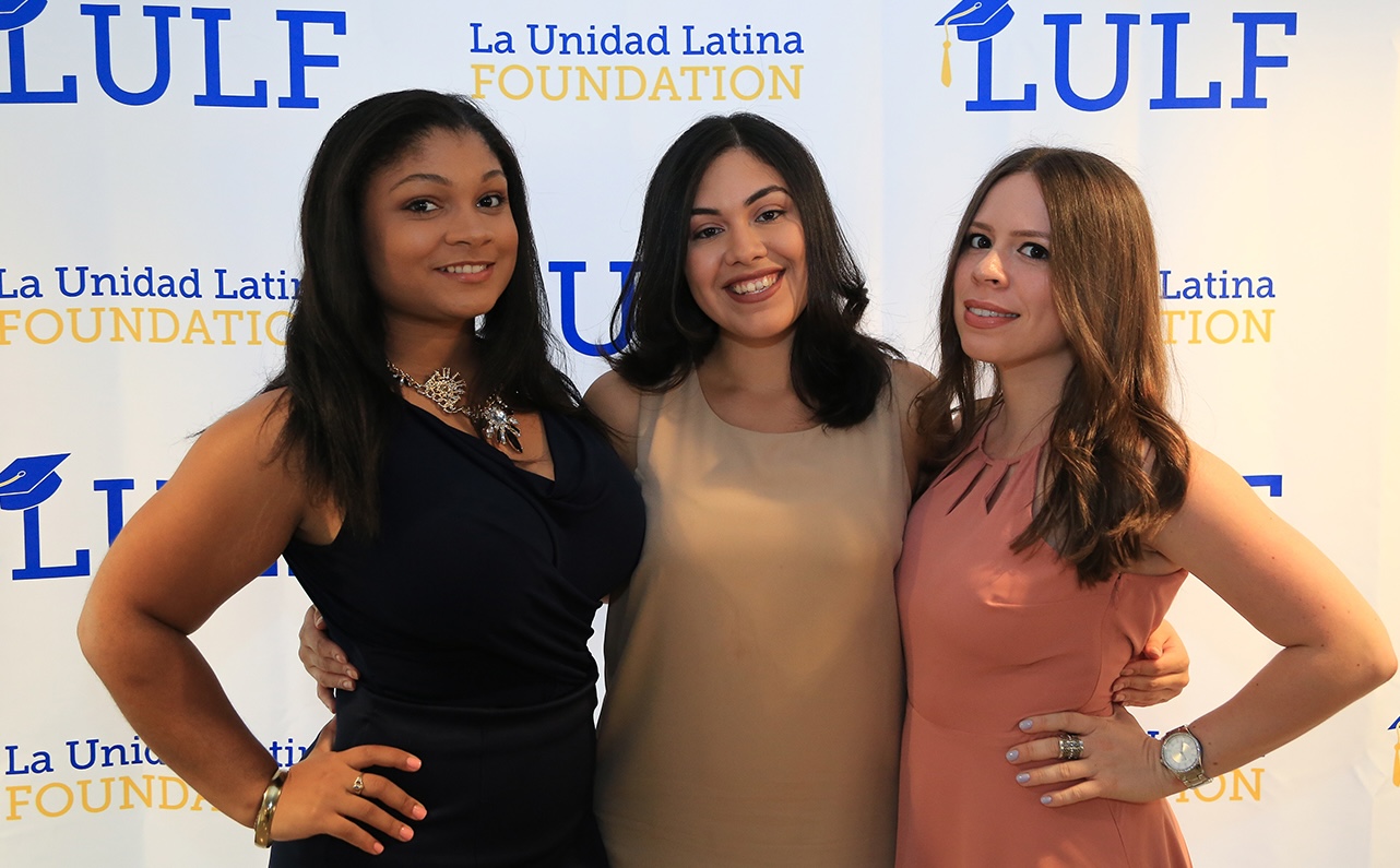 Three women smiling and posing together in front of a La Unidad Latina Foundation backdrop.