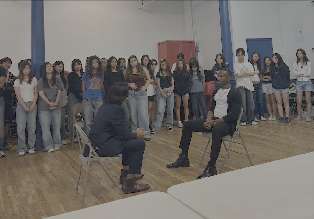 A diverse group of young people standing in a room while two adults sit on chairs in front of them engaged in conversation.