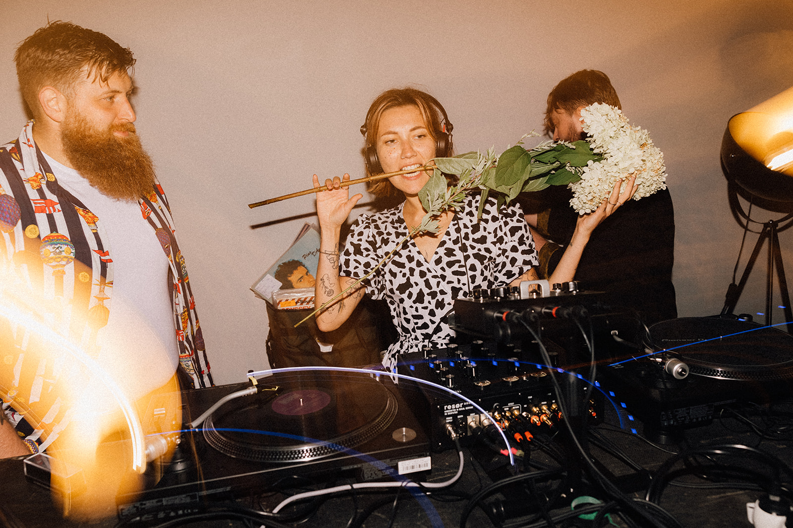 Kobieta-DJ trzyma bukiet w ustach przy konsoli DJ-skiej, obok brodaty mężczyzna w kolorowej koszuli.