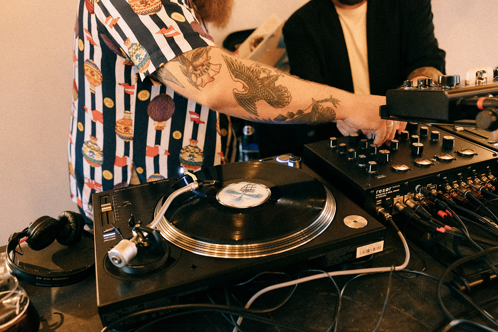 Zbliżenie na konsolę: gramofon, mikser i kable; tatuowana ręka reguluje ustawienia.