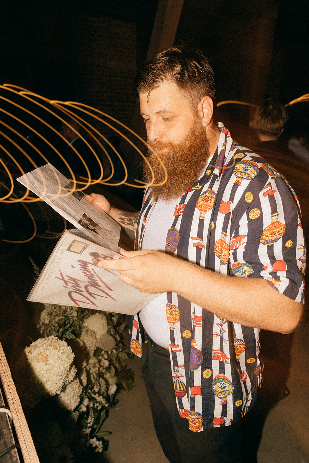 Brodaty mężczyzna w kolorowej koszuli ogląda okładkę winyla „Dirty Dancing”, w tle rozmazane światła i dekoracje kwiatowe.