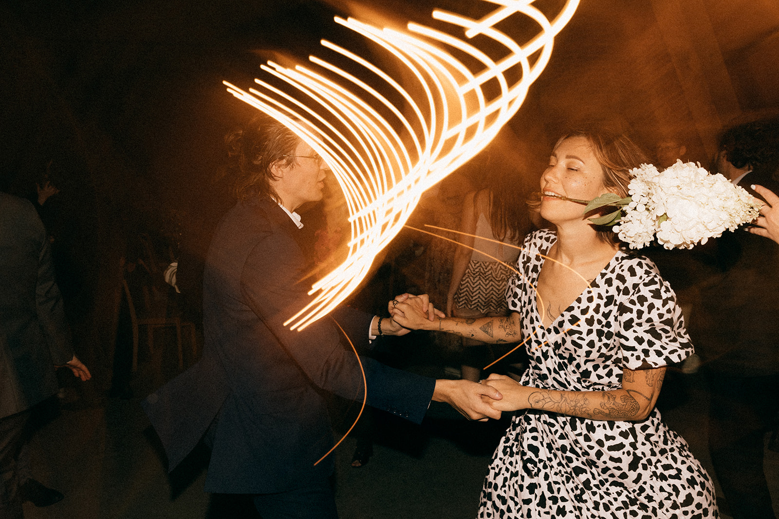 Para tańczy na parkiecie, kobieta trzyma bukiet białych kwiatów w zębach, długie smugi światła w tle.