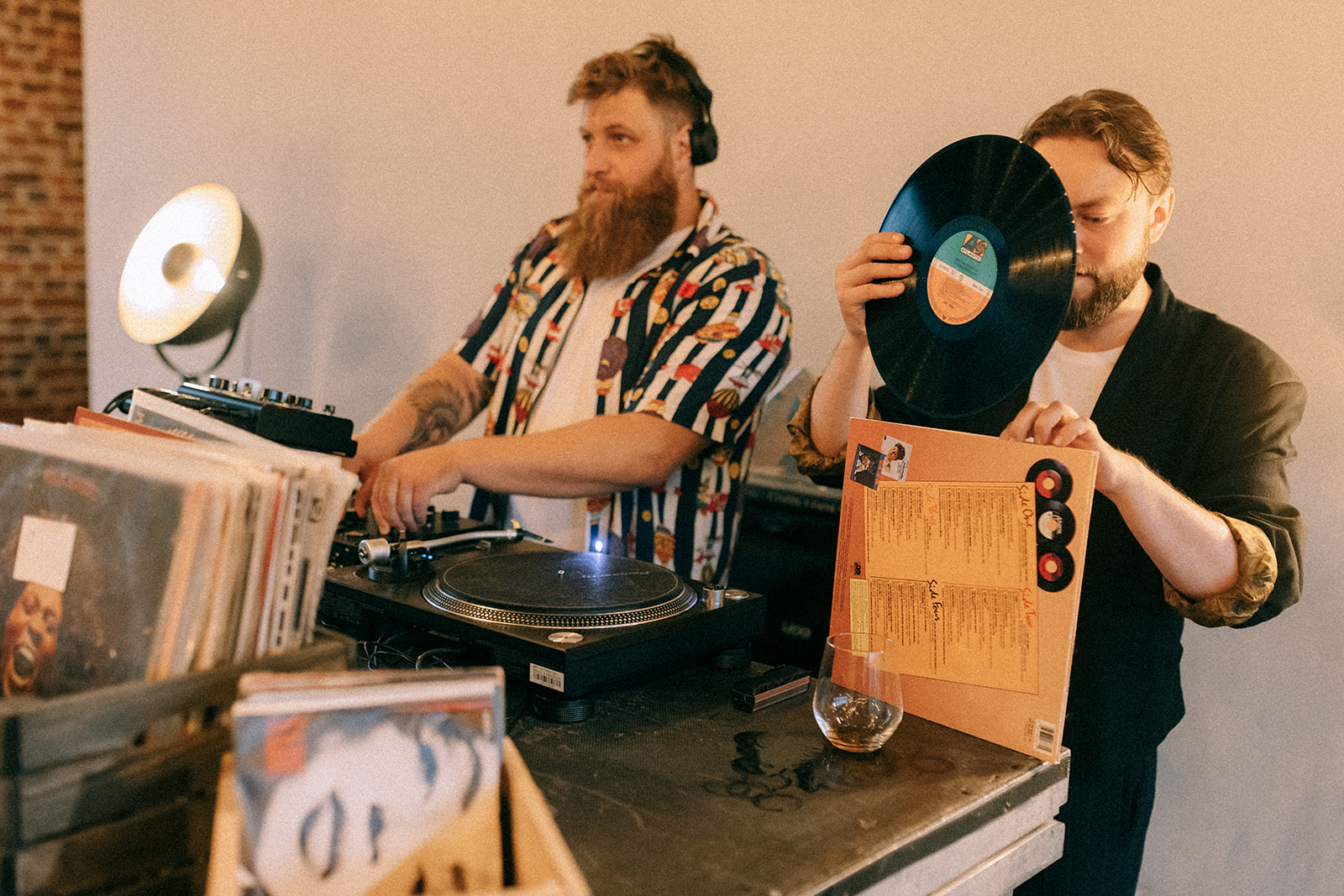 Dwóch DJ-ów za konsoletą: jeden w słuchawkach miksuje, drugi ogląda winyl trzymając okładkę; na blacie szklanka z drinkiem.